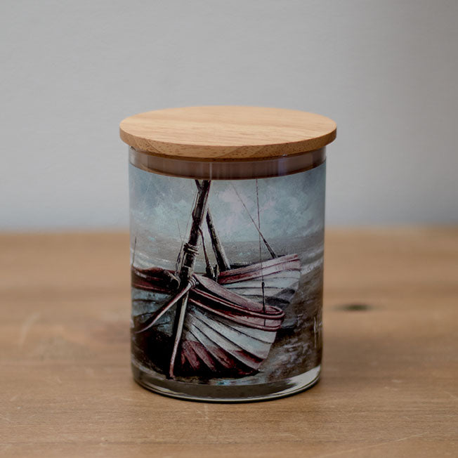 A vanilla scented wax candle with a cotton wick, housed in a clear glass jar featuring a printed image of a boat, with a wooden lid.