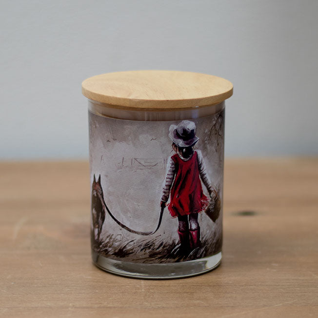A vanilla scented wax candle with a cotton wick, placed in a clear glass jar with a wooden lid, featuring an illustration of a young girl and a dog on the jar's surface.