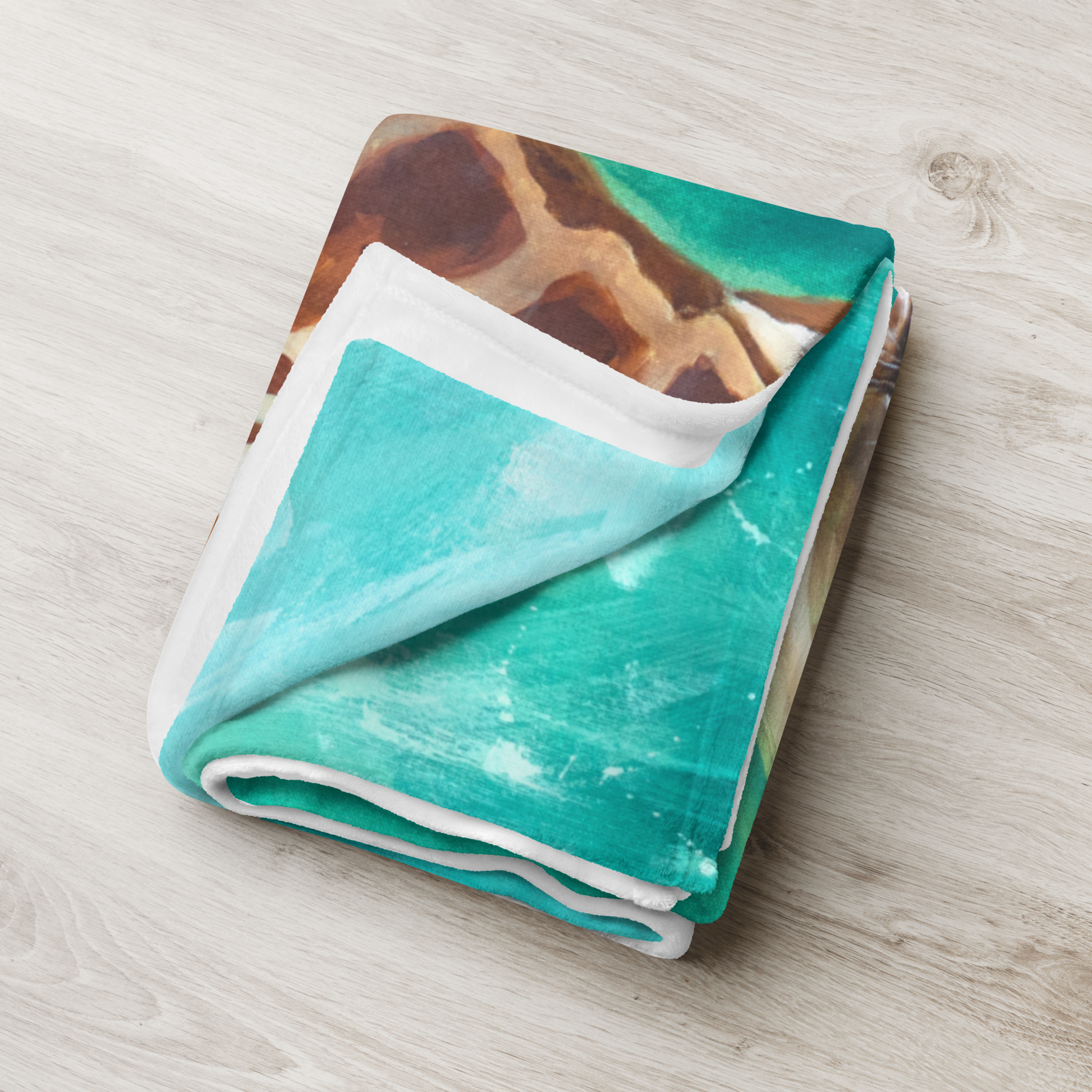 Folded blanket with giraffe pattern on one side and turquoise pattern on the other, on a light wooden surface.