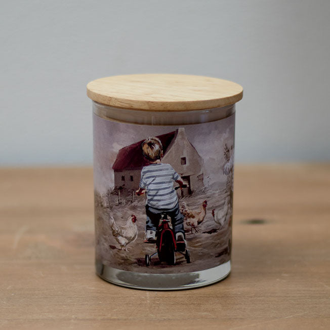 A vanilla scented wax candle with a cotton wick, placed in a clear glass jar with a wooden lid, featuring an illustration of a child on a bicycle.