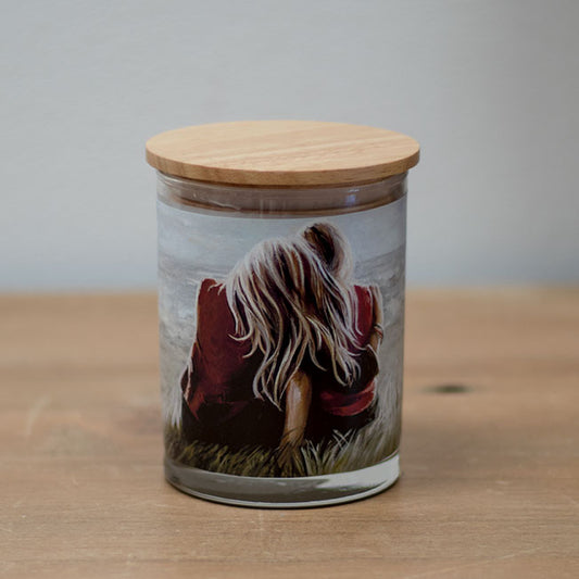 A vanilla scented wax candle with a cotton wick, housed in a clear glass jar with a wooden lid. The jar features a picture of a person sitting with their back towards the viewer, wearing a red coat and facing a grassy landscape.