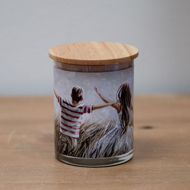 A vanilla scented wax candle with a cotton wick, placed in a clear whisky glass jar with a wooden lid. The jar features an image of two individuals from behind, looking out over a landscape.
