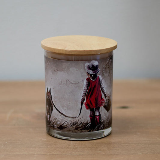 A vanilla scented wax candle with a cotton wick, placed in a clear glass jar with a wooden lid, featuring an illustration of a young girl and a dog on the jar's surface.